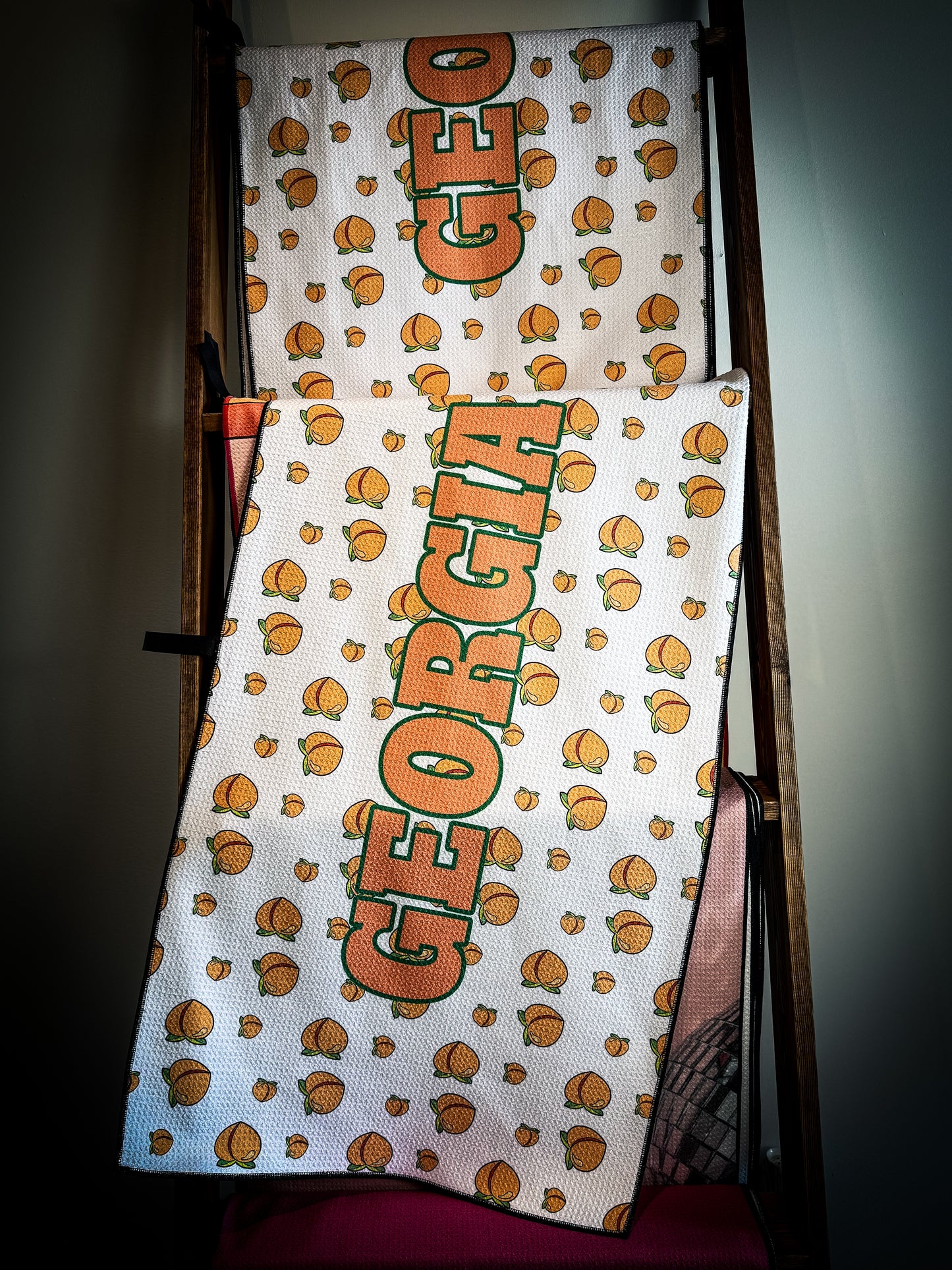 A golf club Towel featuring a pattern with the text 'Georgia' and images of peaches, displayed in a pro shop.